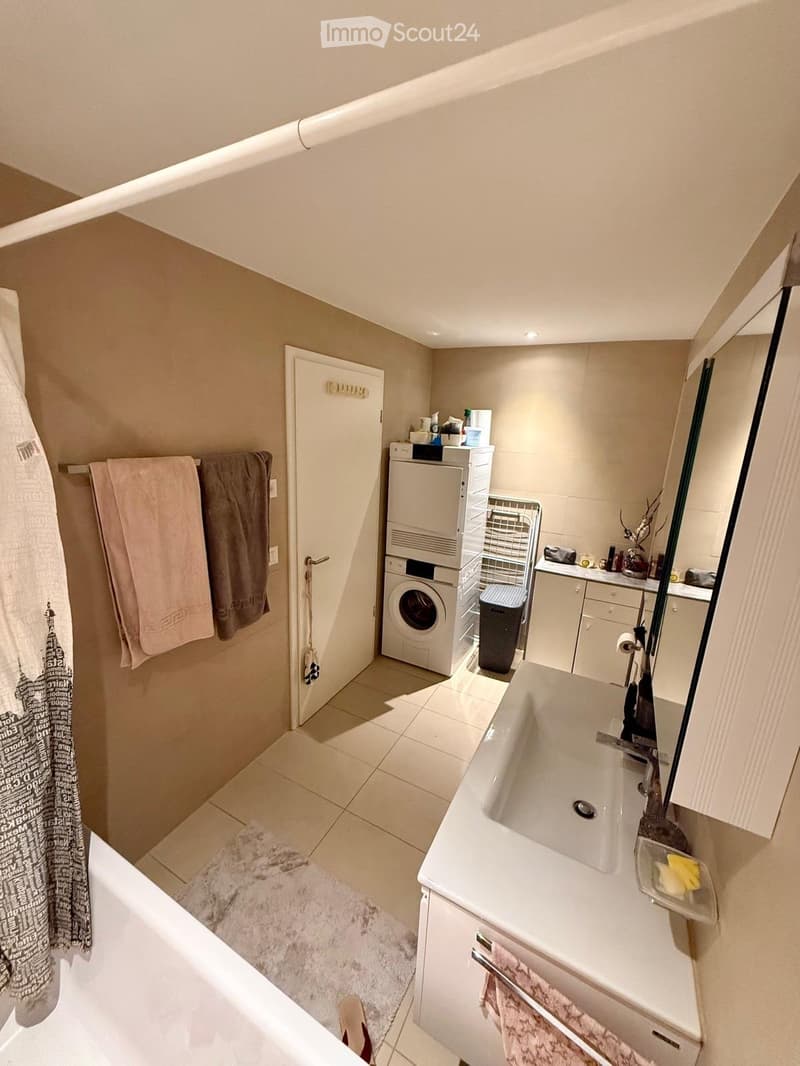 Modern bathroom with a sink, mirror, washing machine, and bathtub. Features tiled floors, white cabinets, and a door leading to another room.
