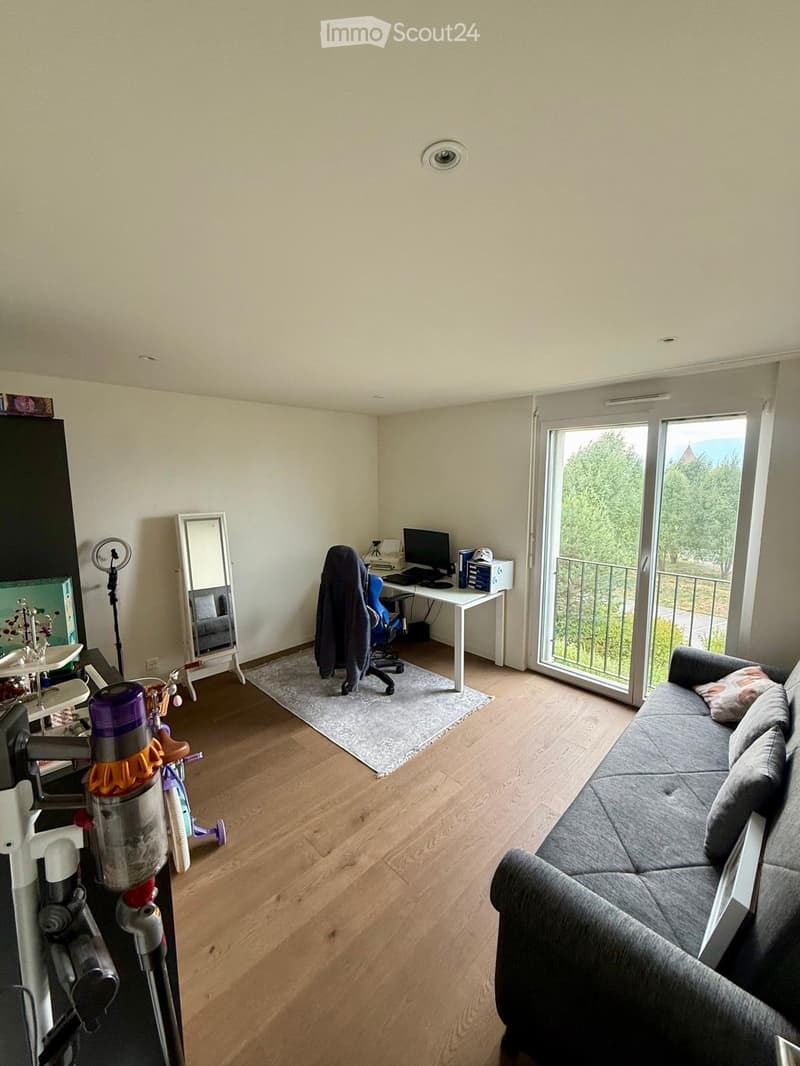A modern living room with a grey sofa, a small table with a computer, a chair, a mirror, and a vacuum cleaner. The room has wooden flooring and large windows with a view of greenery.