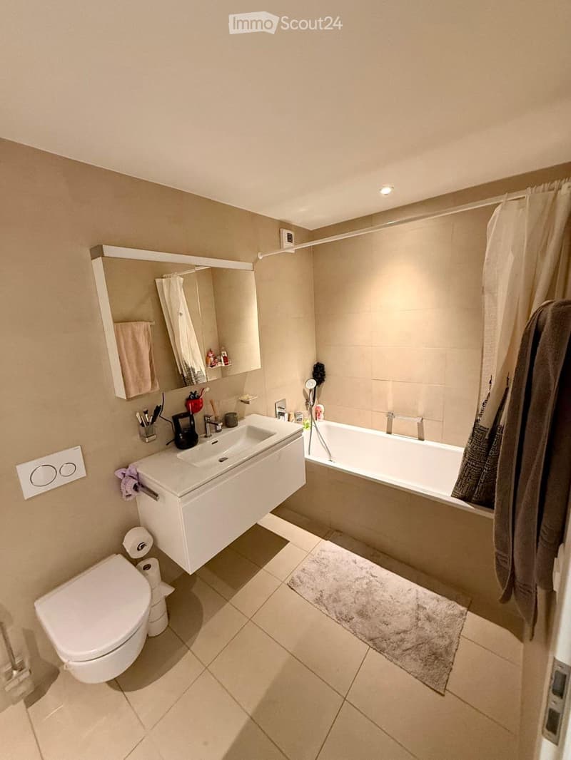 Modern bathroom with beige tiles, a white sink, a bathtub, a toilet, a mirror, and a towel rack. The space includes a soft rug and a bathrobe hanging on the side.