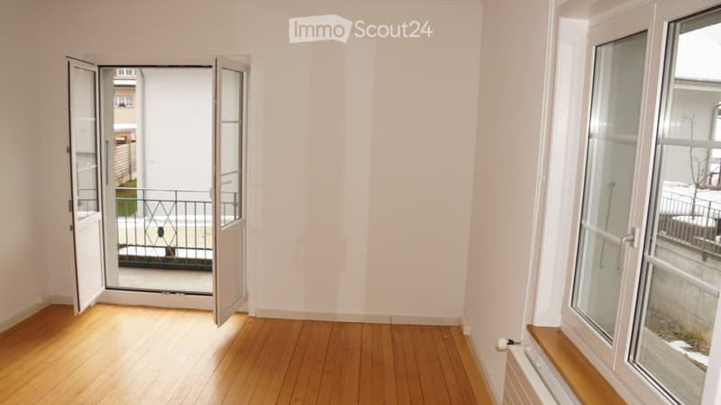 Empty room with wooden flooring, white walls, large windows, and an open door leading to a balcony.