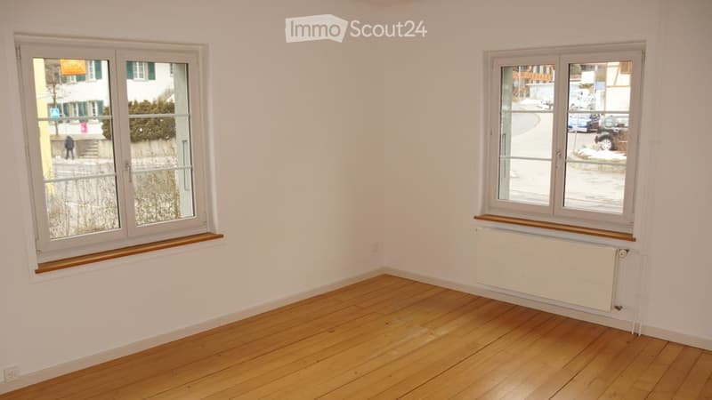 Empty room with wooden flooring, white walls, and two windows with wooden sills. A radiator is visible under one window.