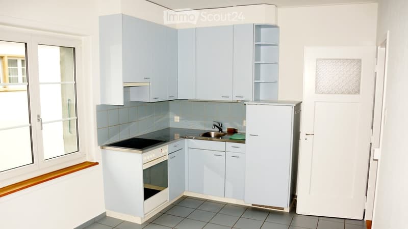 A modern kitchen with light blue cabinets, a white refrigerator, a black electric stove, and a sink with a faucet. The kitchen features tiled walls and a tiled floor, with a window providing natural light.