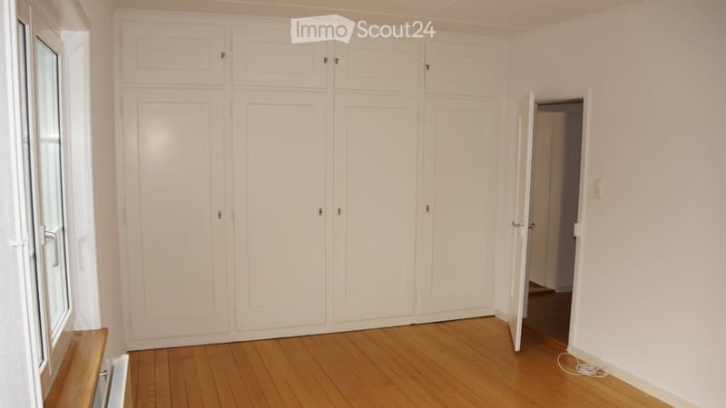Empty room with light-colored wooden flooring, white walls, large built-in wardrobes, and a window with a small bench.