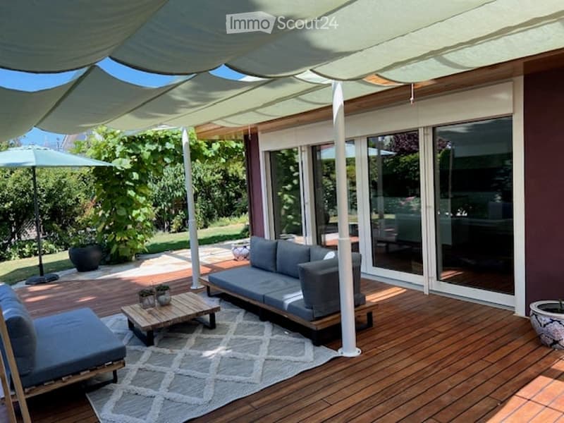 A modern outdoor terrace with wooden decking, grey sofa, coffee table, and a large umbrella providing shade. The space is connected to a house with large glass sliding doors.