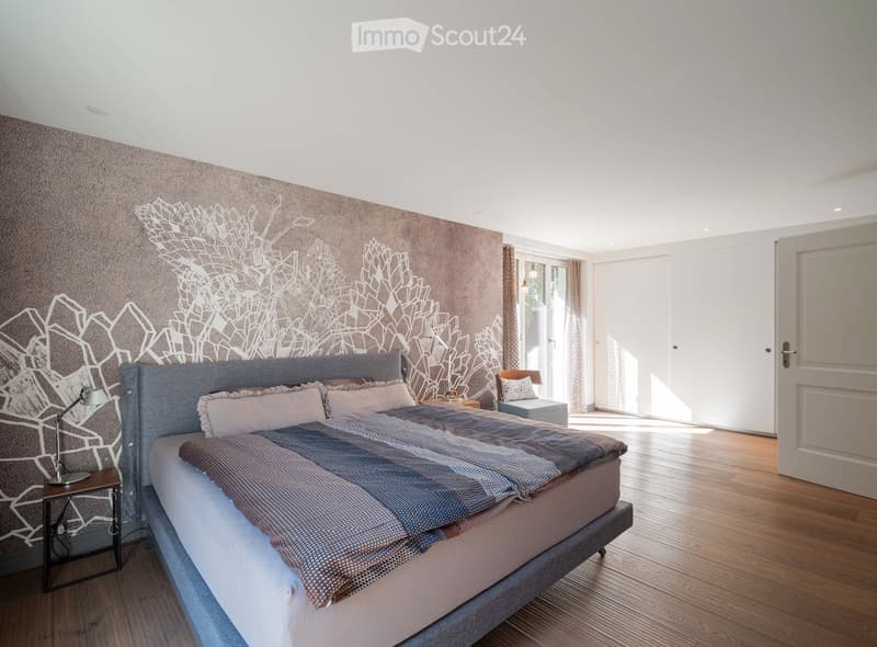 Modern bedroom with a large bed, nightstand, lamp, and a stylish chair near a large window with curtains.