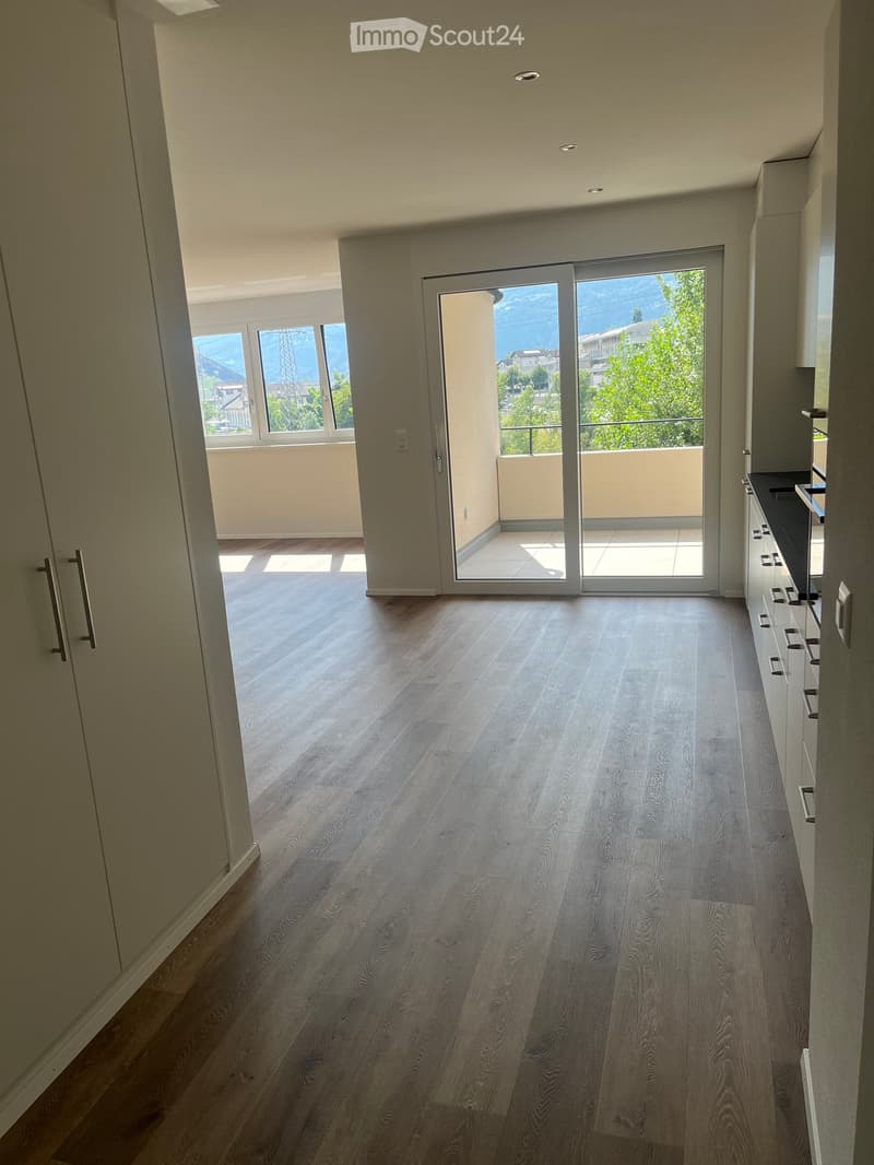 Empty room with wooden flooring, white walls, and large windows offering a view of the outdoors. A sliding glass door leads to a balcony.