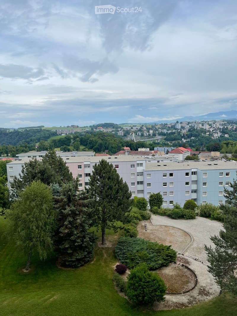 A multi-story residential building complex surrounded by trees and greenery, with a gravel pathway and a view of a cityscape and distant mountains.