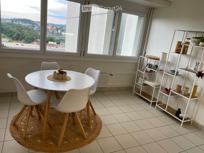 Dining area with a round white table, four white chairs, and a white shelving unit with decorative items. The room has large windows offering a scenic view.