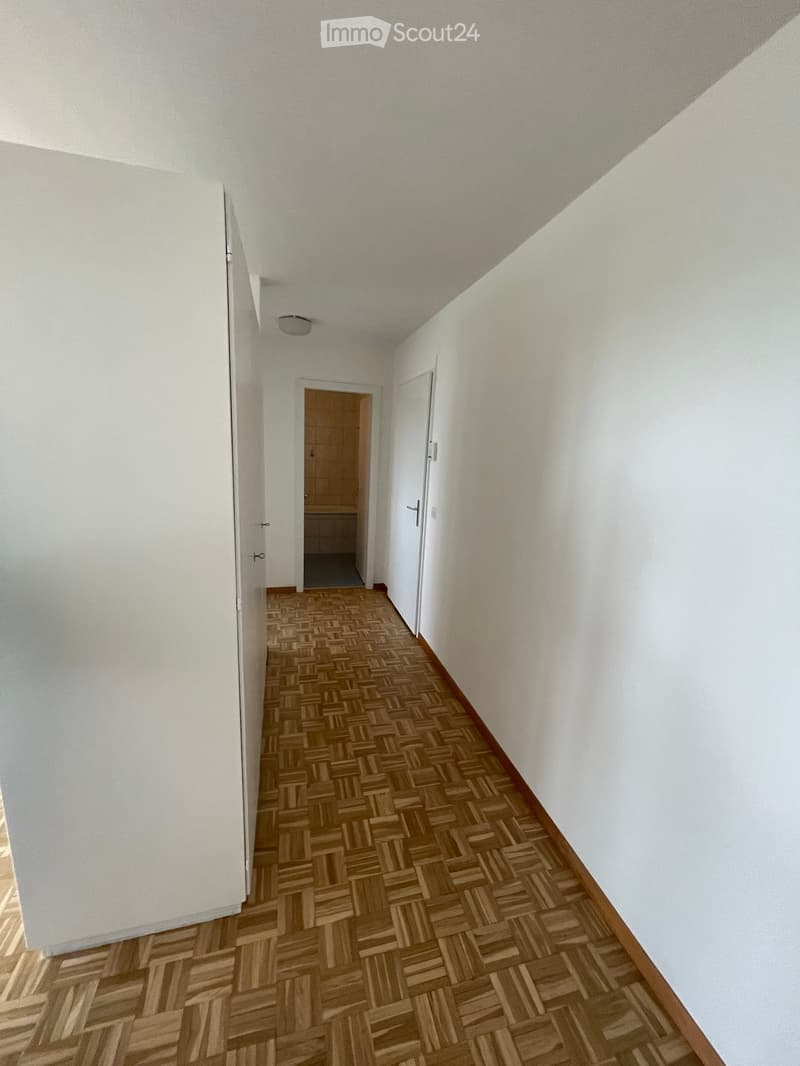 Empty hallway with wooden herringbone flooring, white walls, and a visible bathroom entrance.