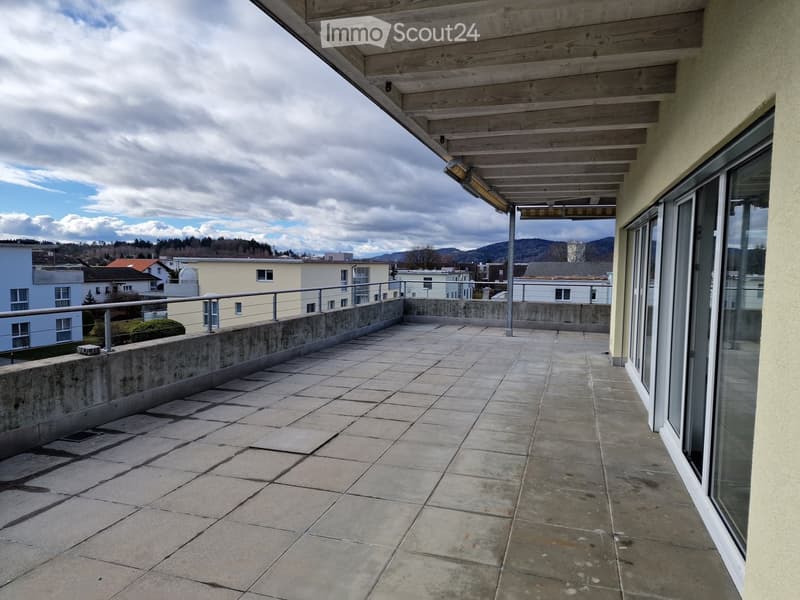An empty balcony with concrete tiles, metal railings, and a glass door on the right side. In the distance, there are buildings, trees, and mountains.