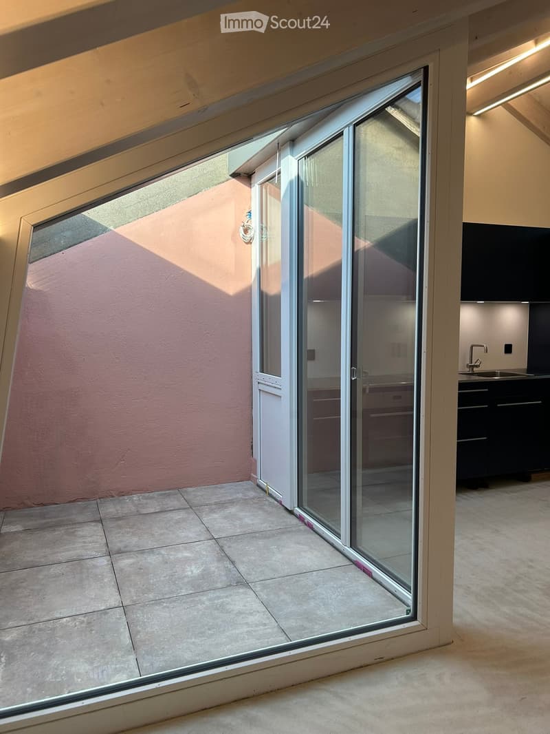 Balcony with large sliding glass doors, tiled floor, and view of a pink wall and adjacent room with kitchen cabinets and sink.