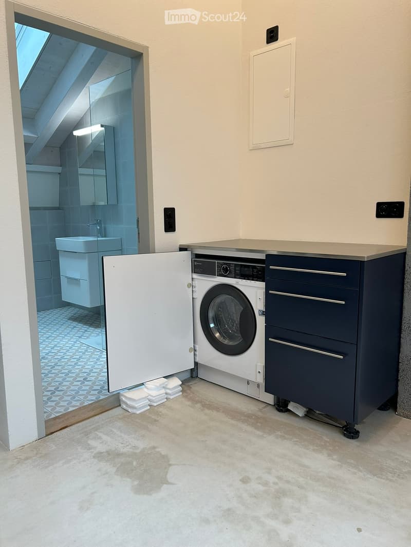 Modern bathroom with a washing machine, sink, mirror, and dark blue cabinet. The floor is concrete, and the walls are light-colored.