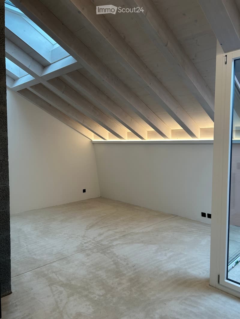 Empty room with wooden floor, white walls, and exposed wooden beams on the ceiling. Natural light streams in from skylights.