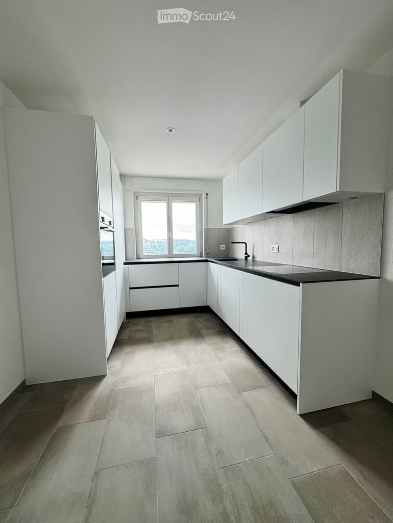 Modern, unfurnished kitchen with white cabinets, a black countertop, and a large window offering a view. The floor is tiled in a neutral tone.