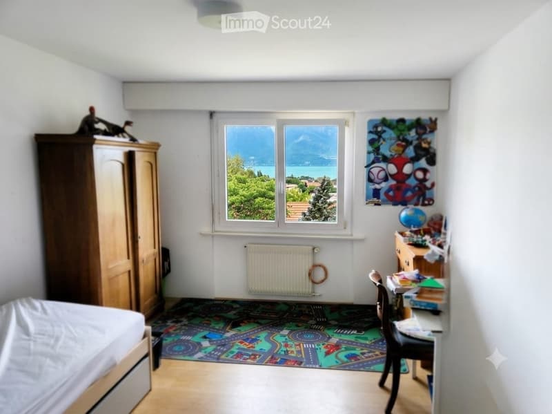 Bright bedroom with large windows offering a scenic view, featuring a bed, wardrobe, desk, chair, globe, and colorful play mat.