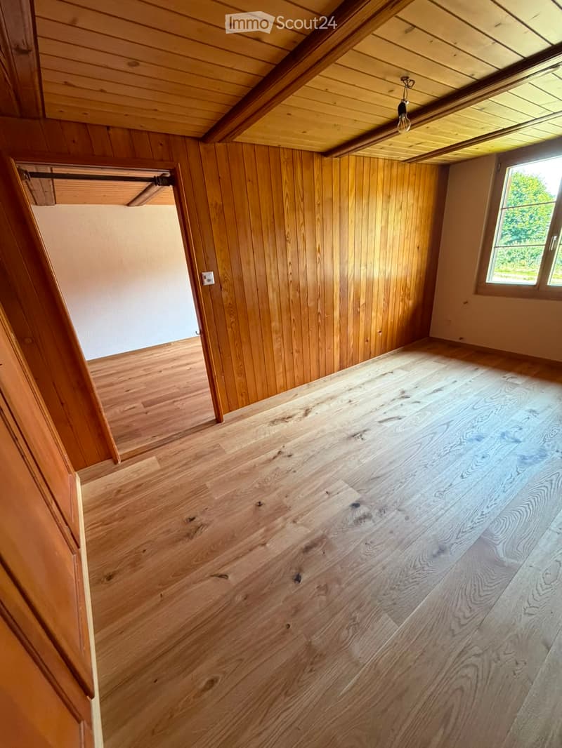 Empty room with wooden walls, wooden floor, and a window letting in natural light. The ceiling has exposed wooden beams and a hanging light fixture.