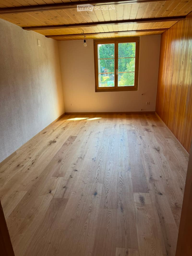 Empty room with wooden flooring, light-colored walls, and a large window with wooden frame. The ceiling is wooden with visible beams and a single hanging light fixture.