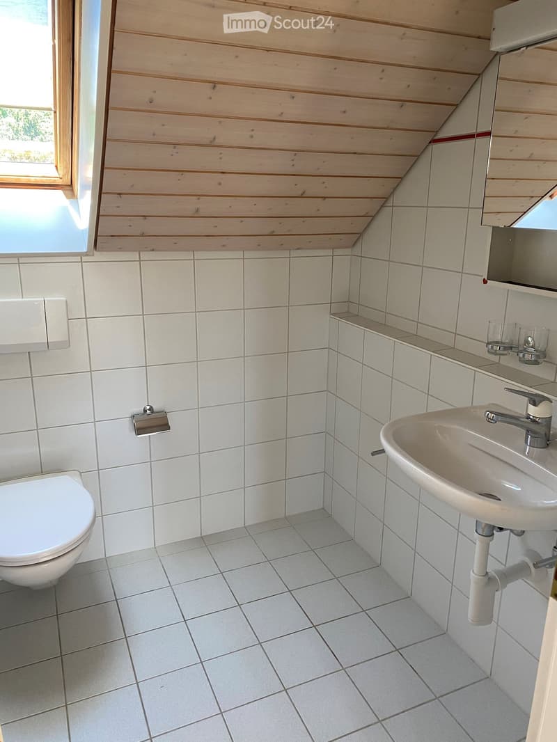 Small bathroom with white tiled walls and floor, a toilet, a sink with a faucet, a mirror, and two cups on the shelf.