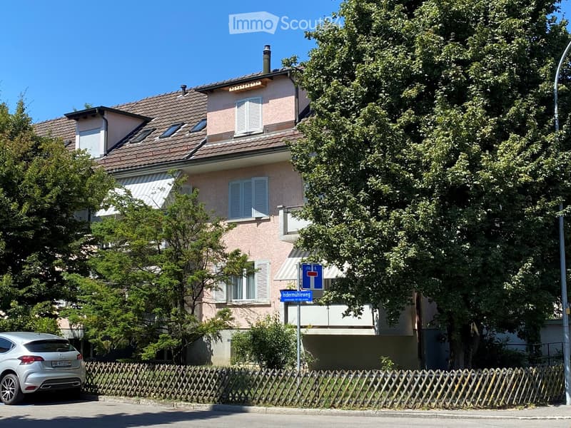Two-story house with a tiled roof, multiple windows with shutters, and a balcony on the upper floor. The house is surrounded by trees and has a wooden fence in front.