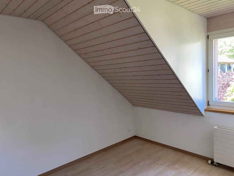 Empty attic room with wooden slatted ceiling, white walls, wooden floor, and a window with a wooden sill. A white radiator is visible on the right side.