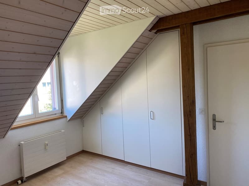 Empty attic room with wooden slanted ceiling, white built-in wardrobes, large window, radiator, and wooden floor.