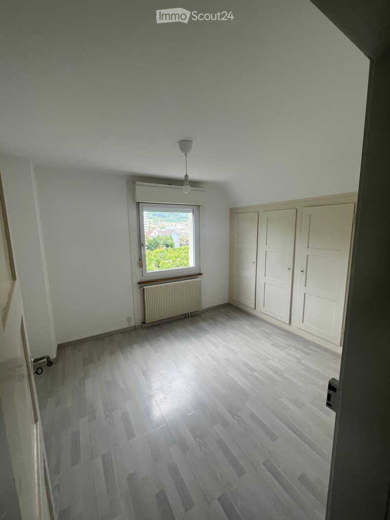 Empty room with light-colored wooden flooring, white walls, and a single window with a radiator below it. The room has white closet doors and a ceiling light fixture.