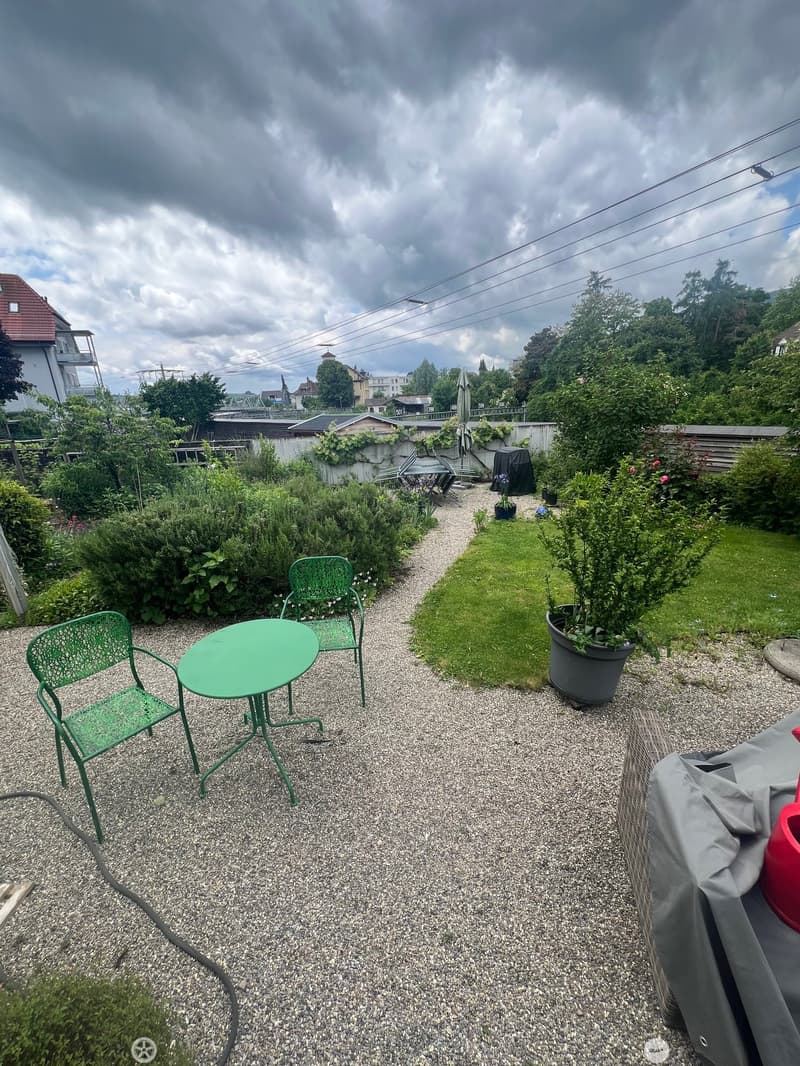 Garden area with gravel path, green metal table and chairs, potted plants, covered grill, and surrounding greenery.