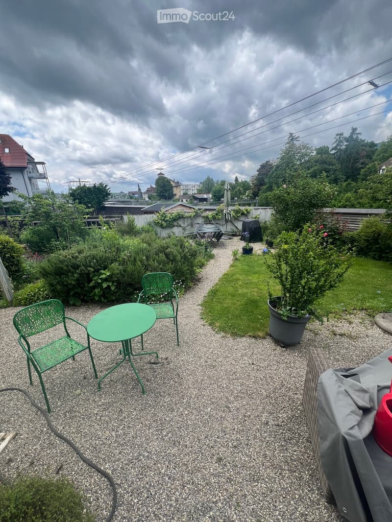 Garden area with gravel path, green metal table and chairs, potted plants, covered grill, and surrounding greenery.