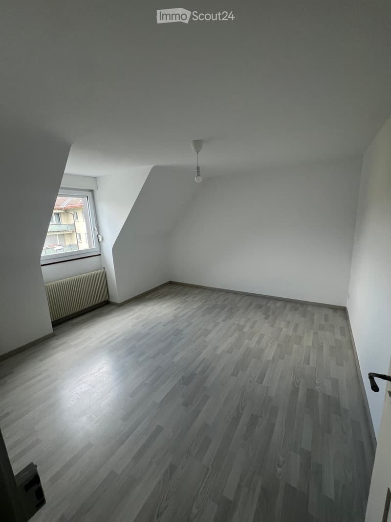 Empty room with wooden flooring, white walls, and a window with a radiator below it. The room has a slanted ceiling and a single light fixture hanging from the ceiling.