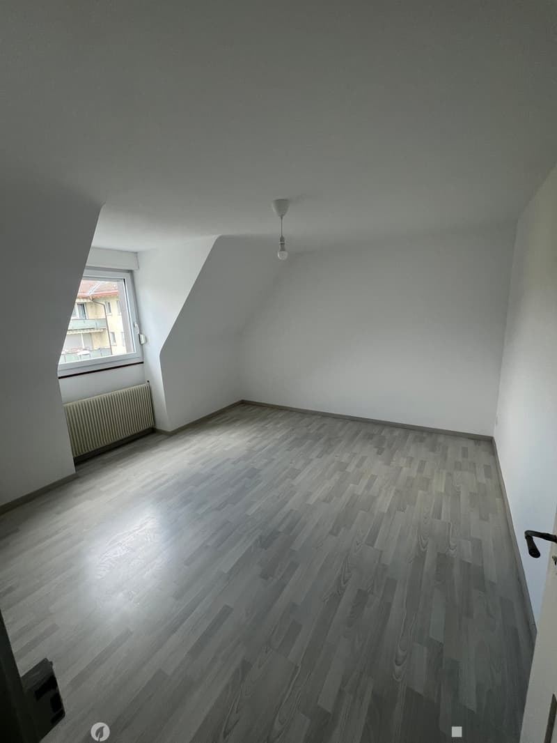 Empty room with wooden flooring, white walls, and a window with a radiator below it. The room has a slanted ceiling and a single light fixture hanging from the ceiling.