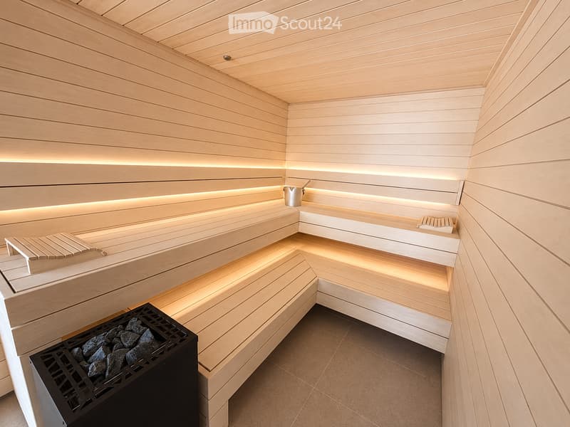 Modern sauna with wooden benches, integrated lighting, stone rocks in a black container, and a metal bucket on the upper bench.
