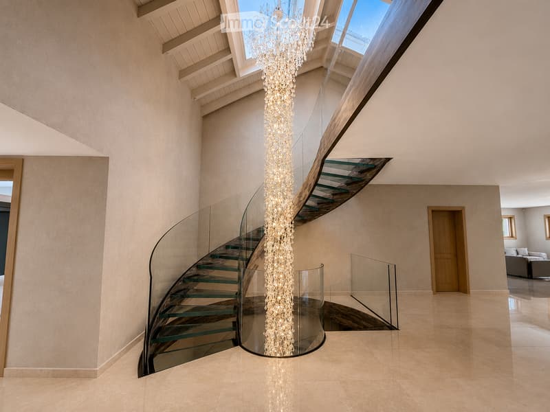 Spacious hallway with a modern glass staircase, a tall decorative light fixture, and a skylight above. The walls are light-colored, and the floor is tiled.