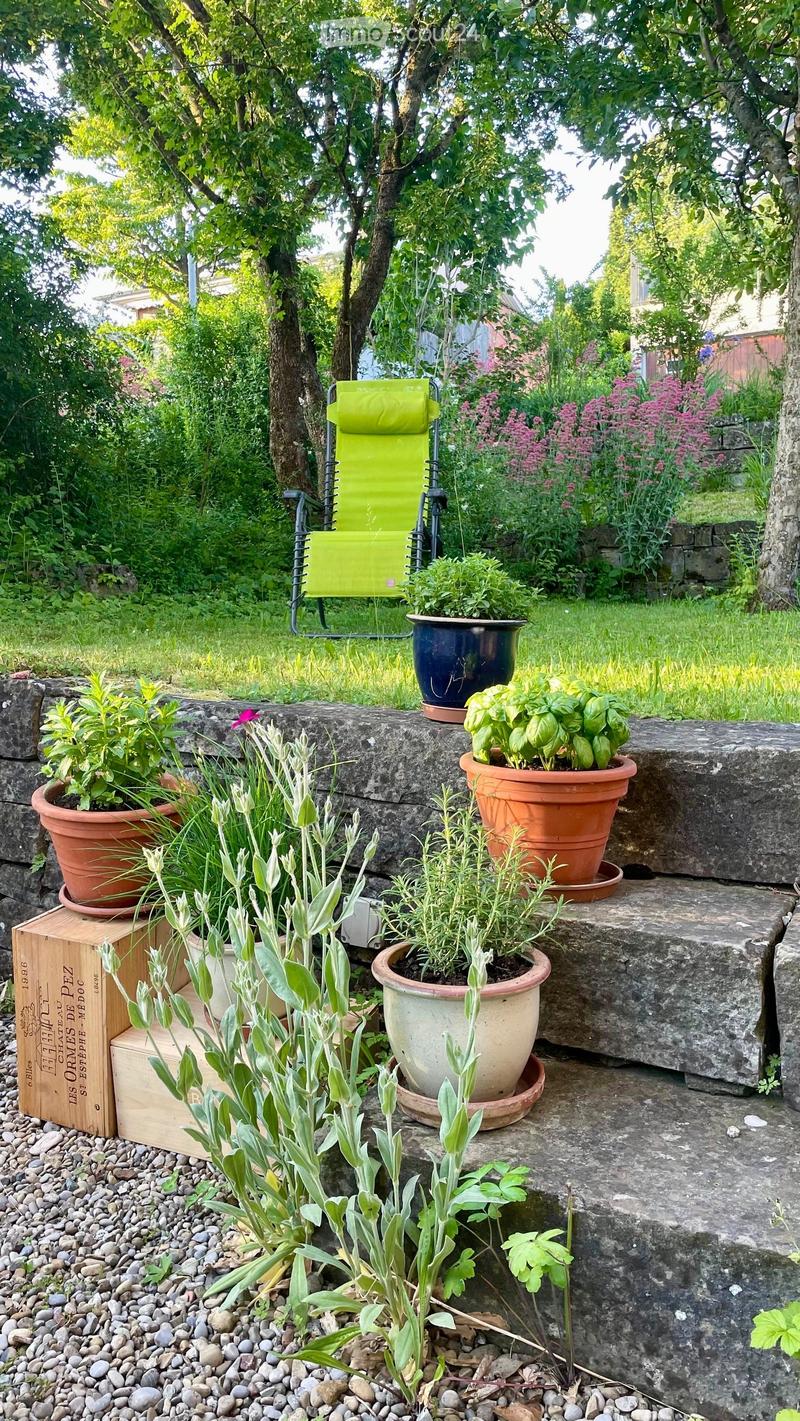 A lush garden with potted plants, a green folding chair, and a stone wall. The garden features various plants in terracotta pots, a wooden crate, and a stone pathway.