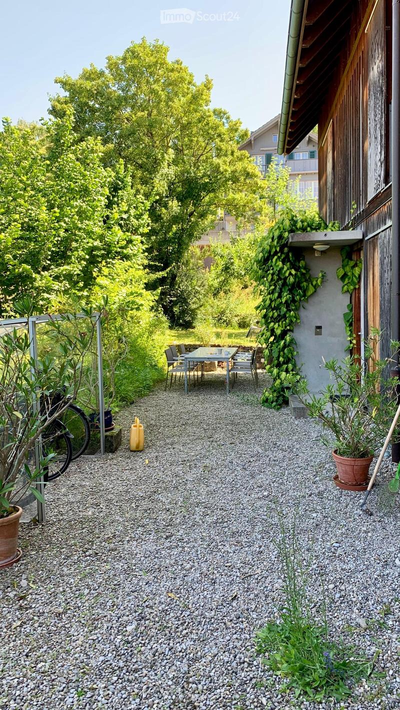 Gravel pathway leading to a small outdoor seating area with a table and chairs, surrounded by greenery and potted plants.