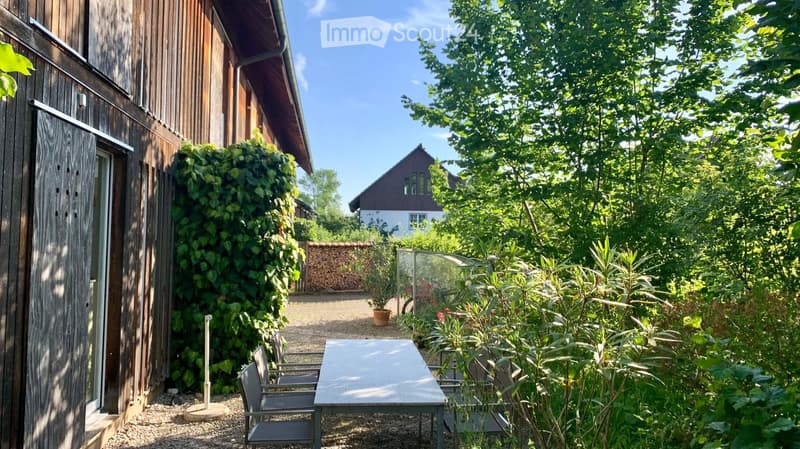 A lush garden area with a wooden house, a patio table and chairs, a potted plant, and a stack of firewood.