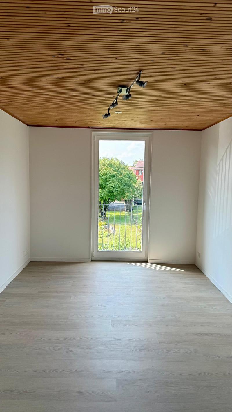 Empty room with light wooden flooring, white walls, and a large glass door leading to a green outdoor area.