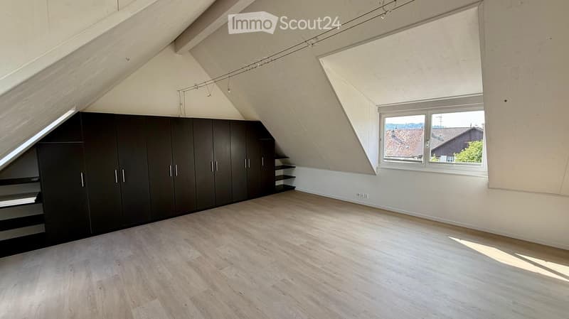 Empty attic room with wooden flooring, built-in dark brown cabinets, and large skylight windows offering a view of the neighborhood.