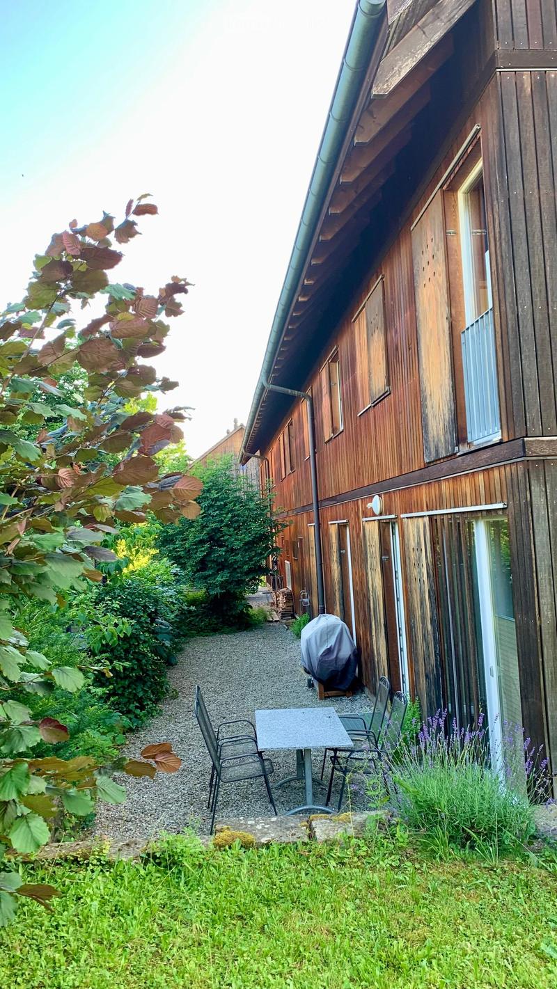 A narrow garden path leads to a wooden building with a covered patio area featuring a table and chairs, surrounded by greenery and lavender plants.