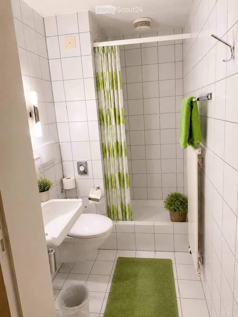 Modern bathroom with white tiled walls and floor, featuring a sink, toilet, bathtub with a green and white shower curtain, a green bath mat, and decorative plants.