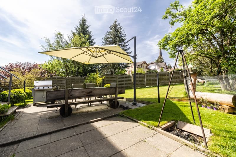 Well-maintained garden with a patio area, outdoor furniture, a large umbrella, a barbecue, and a swing set.