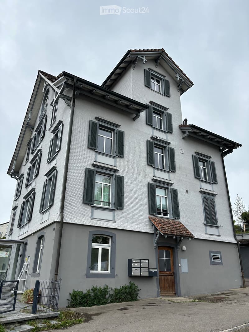 A multi-story residential house with a textured light gray facade, dark green shutters, and multiple windows. The entrance features a wooden door with a small awning, and there is a small garden area with shrubs near the entrance.