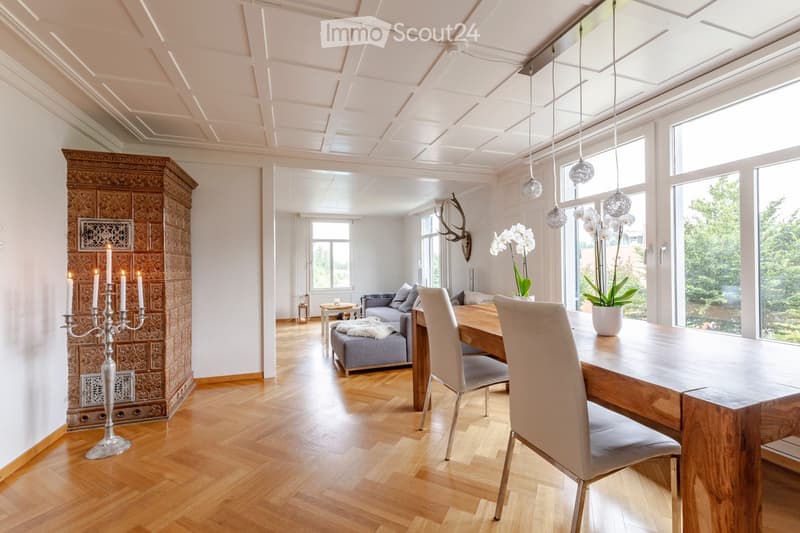 Elegant living and dining area with wooden flooring, a grey sofa with an ottoman, a wooden dining table with chairs, a candelabra, and vases with white flowers. Large windows and a decorative cabinet are also visible.