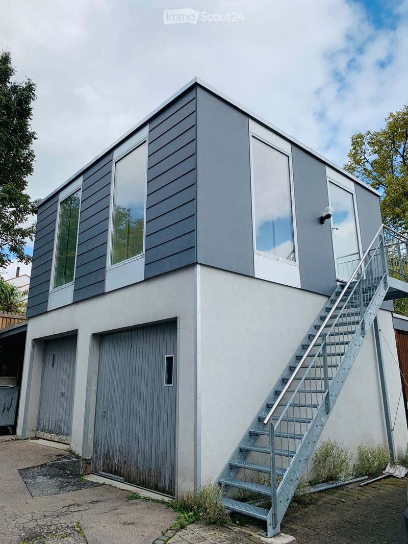 Two-story modern house with grey siding, large windows, raised ground floor, and a metal staircase leading to an upper-level balcony.