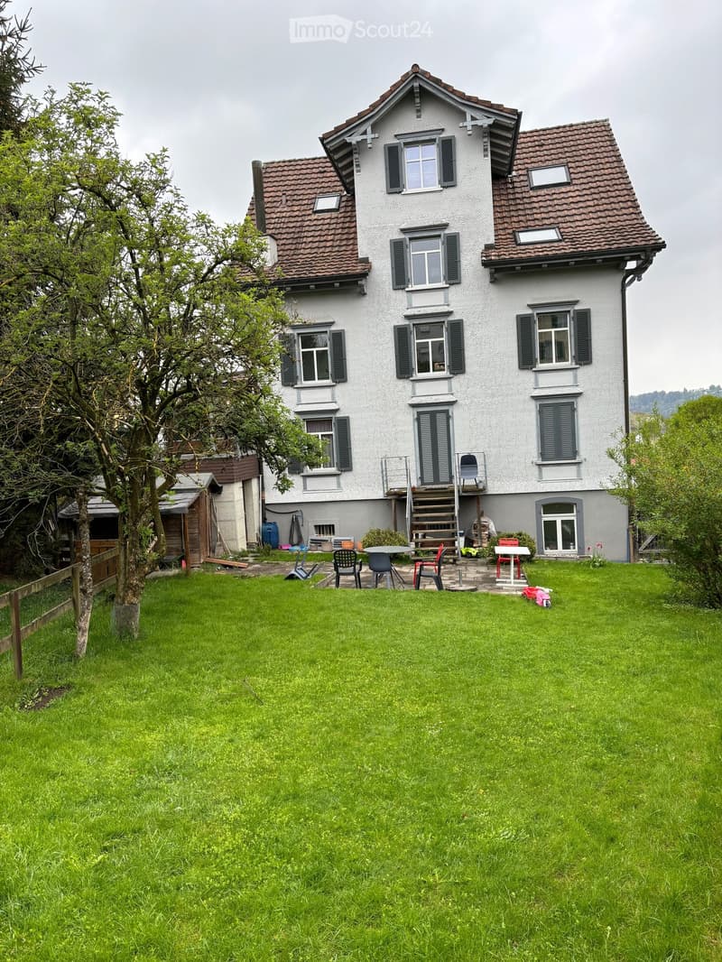 A two-story house with a red-tiled roof and multiple windows, surrounded by a well-maintained lawn and a small patio area with outdoor furniture.