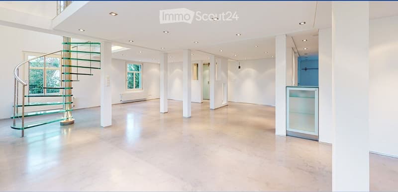 Open, spacious room with a modern glass spiral staircase, large windows, and recessed lighting. The floor is polished concrete, and the walls are white.