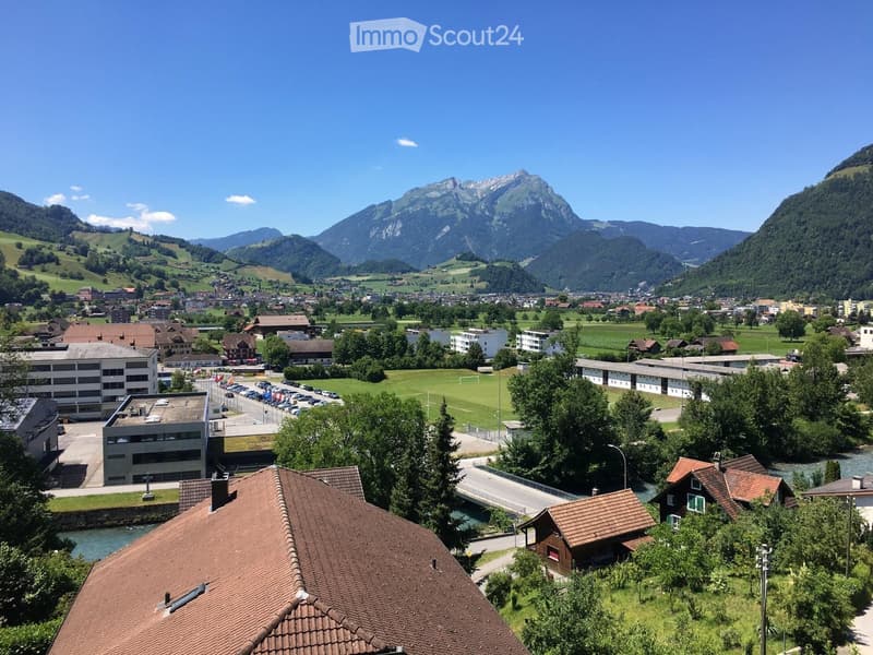 A panoramic view of a town nestled in a valley with surrounding mountains, featuring residential buildings, a sports field, and a river running through the area.
