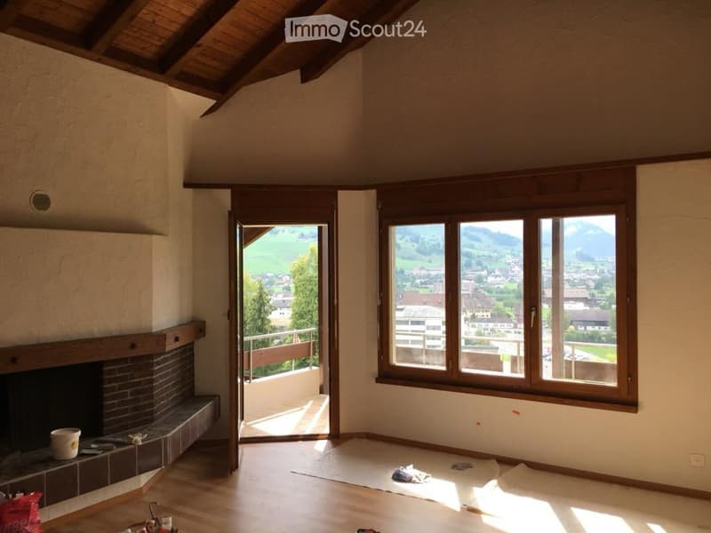 Bright, unfurnished room with wooden floors, exposed wooden beams, large windows offering scenic mountain views, and an open doorway leading to a balcony.