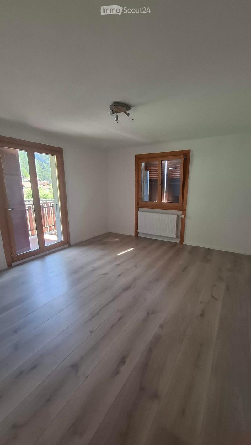 Empty room with wooden flooring, white walls, and a ceiling light fixture. Features a large window with wooden frame and a sliding glass door leading to an outdoor area.