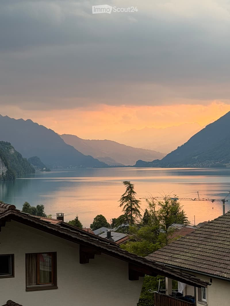 View of a serene lake surrounded by mountains at sunset, with a house in the foreground featuring a tiled roof and visible windows.