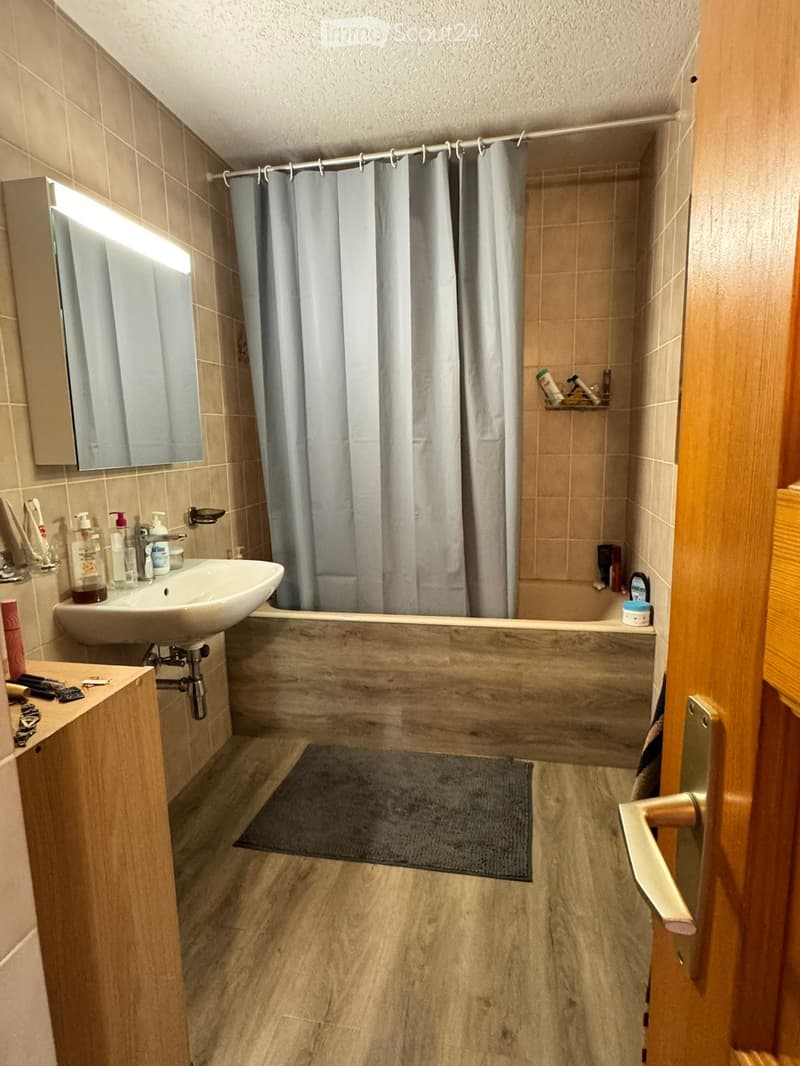 Modern bathroom with a white sink, mirror, bathtub with a shower curtain, and a wooden cabinet. The floor is tiled, and a gray rug is placed in front of the bathtub.